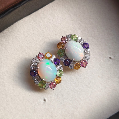 A pair of oval stud earrings with white opal centers surrounded by multicolored gemstones, displayed on a cream-colored jewelry box.
