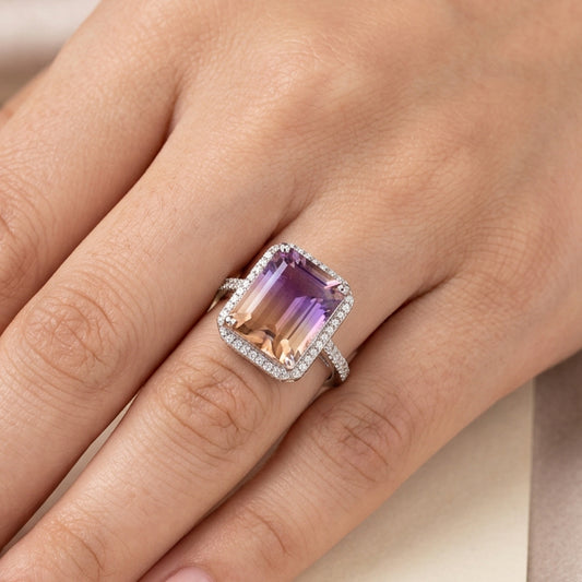A close-up of a hand wearing a silver ring with a large rectangular pinkish-purple gemstone and a halo of small clear stones.