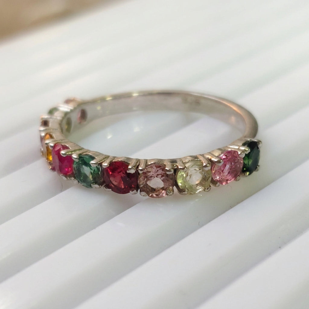 A delicate gold eternity ring featuring multiple small colorful gemstones, including pink, green, and yellow stones, resting on a white ribbed surface.