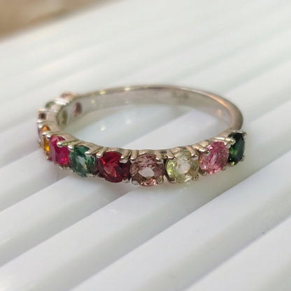 A delicate gold eternity ring featuring multiple small colorful gemstones, including pink, green, and yellow stones, resting on a white ribbed surface.