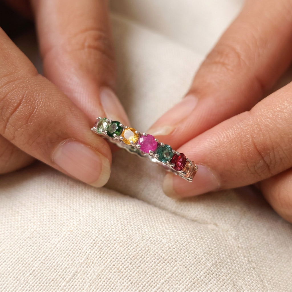 A colorful gemstone eternity ring displayed inside a black jewelry box with a cream-colored lining. A hand with pink nail polish is holding the left side of the box, and a white paper tag is attached.