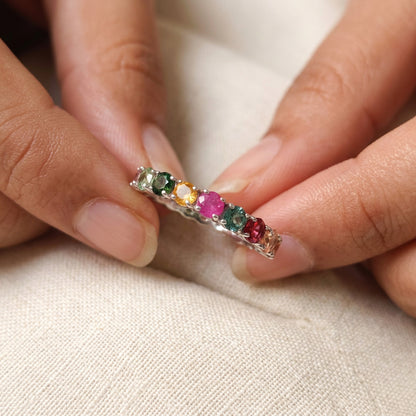 A colorful gemstone eternity ring displayed inside a black jewelry box with a cream-colored lining. A hand with pink nail polish is holding the left side of the box, and a white paper tag is attached.