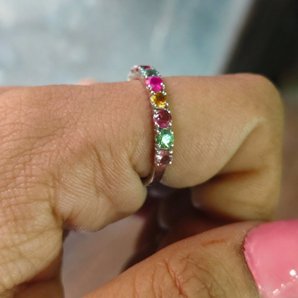 A close-up of a silver band ring with a row of colorful gemstones (pink, yellow, green) worn on a person's finger.
