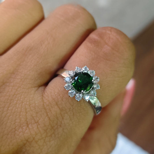 A silver ring with a round green emerald-cut center stone and a halo of small clear stones, shown worn on a person's finger.