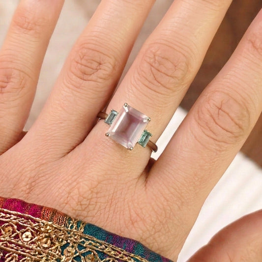 A close-up of a hand wearing a silver ring with a large rectangular pale pink gemstone and two small light blue accent stones on either side of the band.