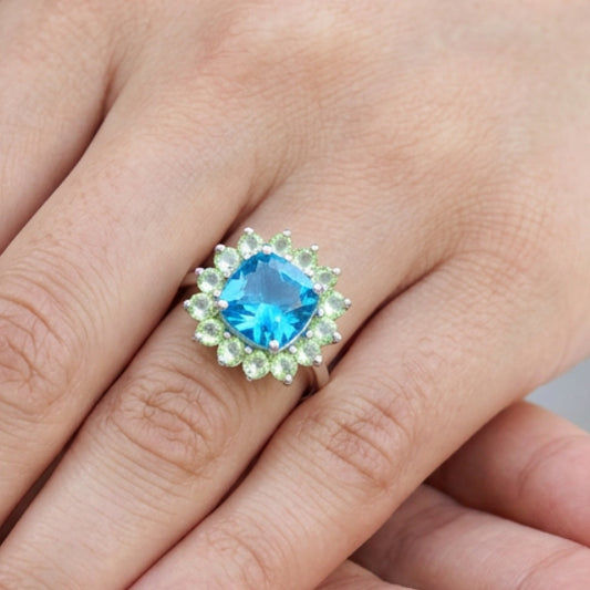 A close-up of a hand wearing a silver ring with a large blue cushion-cut center stone surrounded by a halo of small light green stones.