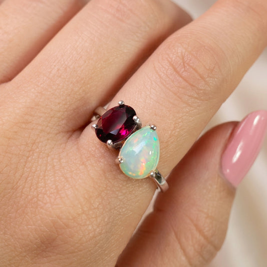 A close-up of a hand wearing a silver ring with a marquise-cut light green opal and a round dark red gemstone.