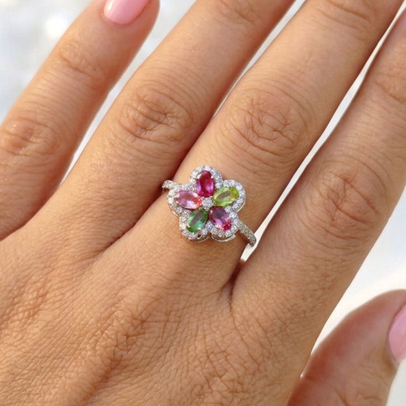 A silver-toned flower ring with four marquise-cut gemstones in pink, green, and purple, worn on a finger. The ring is surrounded by small clear stones, and the background is a soft-focus indoor setting.