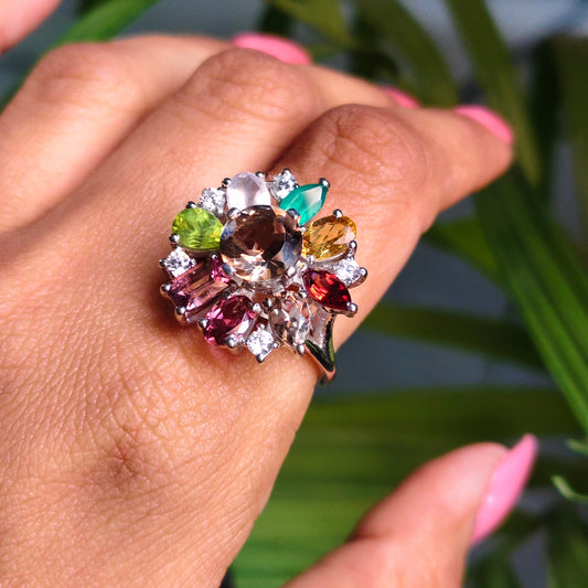 A close-up of a hand with pink manicured nails wearing a large, colorful gemstone cluster ring. The ring features multiple teardrop-cut gemstones in shades of green, pink, purple, orange, and brown on a silver-toned setting, set against a blurred background of green leaves.