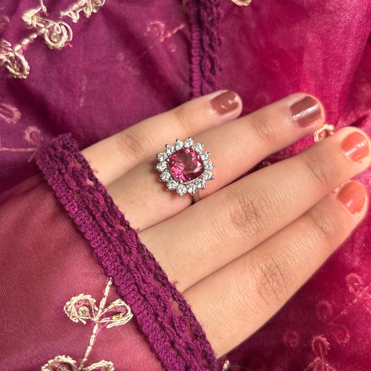 A close-up of a hand wearing a silver ring with a round pink gemstone surrounded by small clear stones, set against a magenta embroidered fabric background.