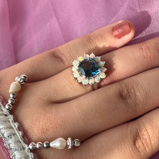 A close-up of a hand wearing a silver ring with a round blue gemstone surrounded by small white stones, styled with beaded bracelets against a pink fabric background.
