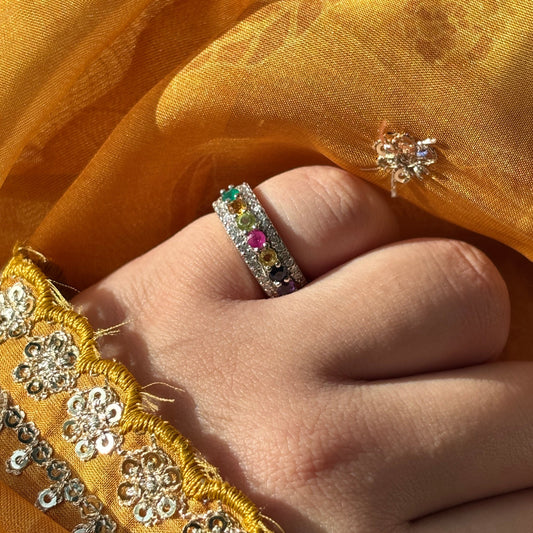 A close-up of a hand wearing a silver ring with colorful gemstones, set against a background of golden-yellow embroidered fabric.