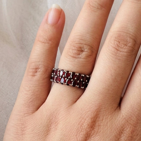 A close-up of a hand wearing a beaded ring with dark red and black beads, resting on a sheer embroidered sleeve against a soft white fabric background.
