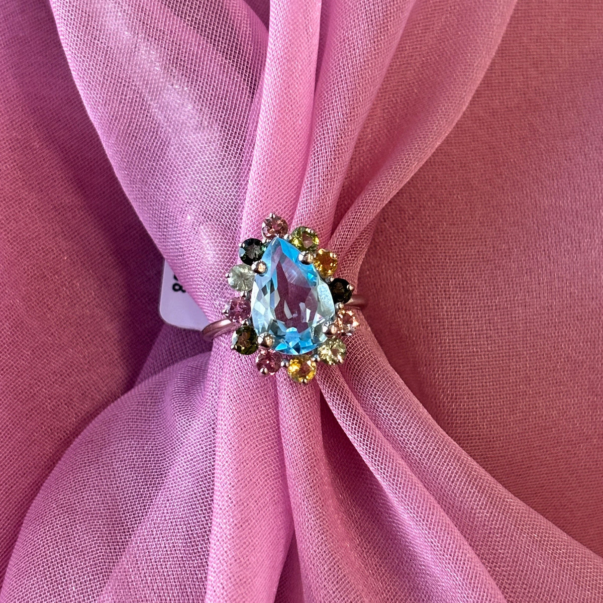 A close-up of a ring with a marquise-cut light blue center stone and small green and yellow accent stones, displayed on pink sheer fabric.