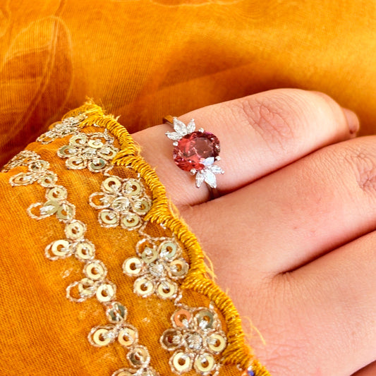 A close-up of a hand wearing a silver ring with a round pink gemstone, resting against a textured golden-yellow embroidered fabric background.