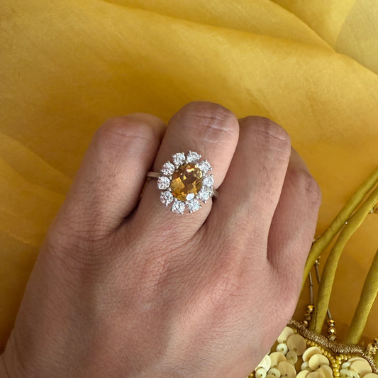 A close-up of a hand wearing a silver ring with a central round yellow gemstone surrounded by small clear stones, set against a bright yellow fabric background.