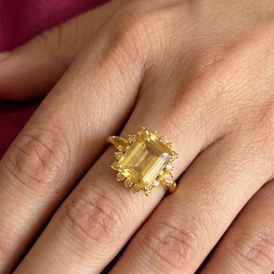 A close-up of a hand wearing a yellow gold ring with a large rectangular yellow gemstone, set against a background of yellow fabric and sheer white fabric.