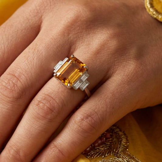 A close-up of a hand wearing a silver ring with a large rectangular amber-yellow gemstone, set against a background of white fabric and a yellow embroidered sleeve.