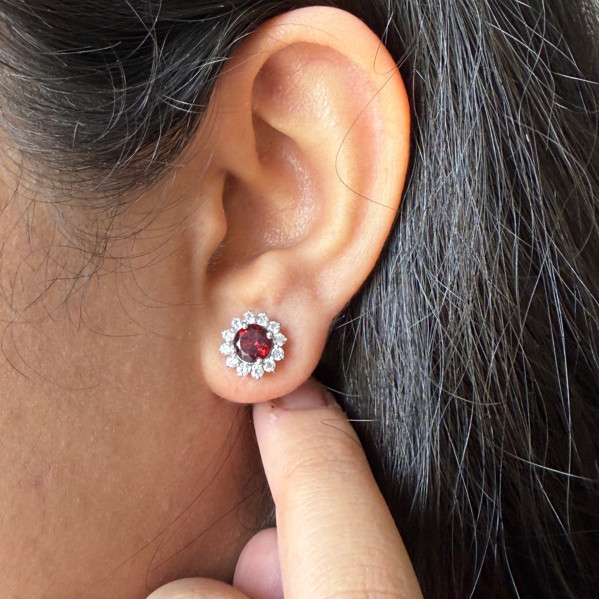 A close-up of a person's ear wearing a small stud earring with a dark red center stone surrounded by a halo of clear stones, with their finger touching the lobe below the earring.