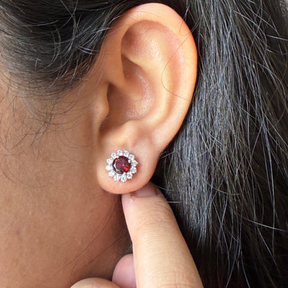 A close-up of a person's ear wearing a small stud earring with a dark red center stone surrounded by a halo of clear stones, with their finger touching the lobe below the earring.