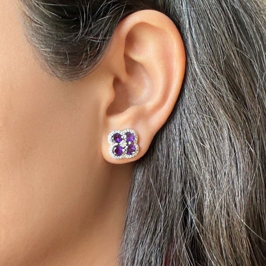 A close-up of a person's ear wearing a small four-stone stud earring with purple gemstones surrounded by clear stones in a silver-toned setting.