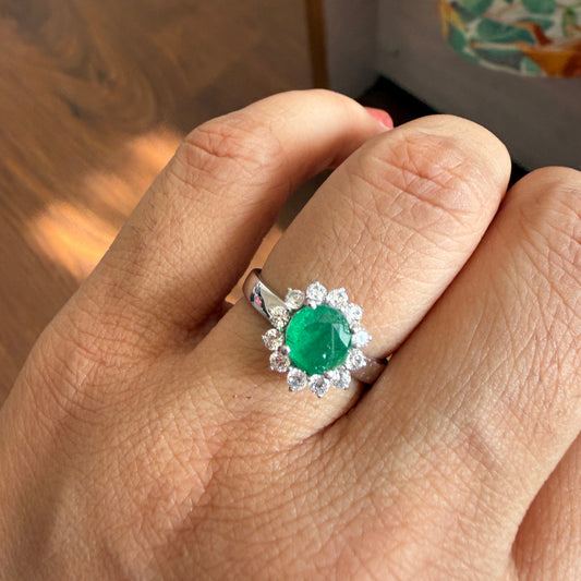 A silver ring featuring a round green emerald-cut center stone surrounded by a halo of small clear stones, worn on a person's finger.