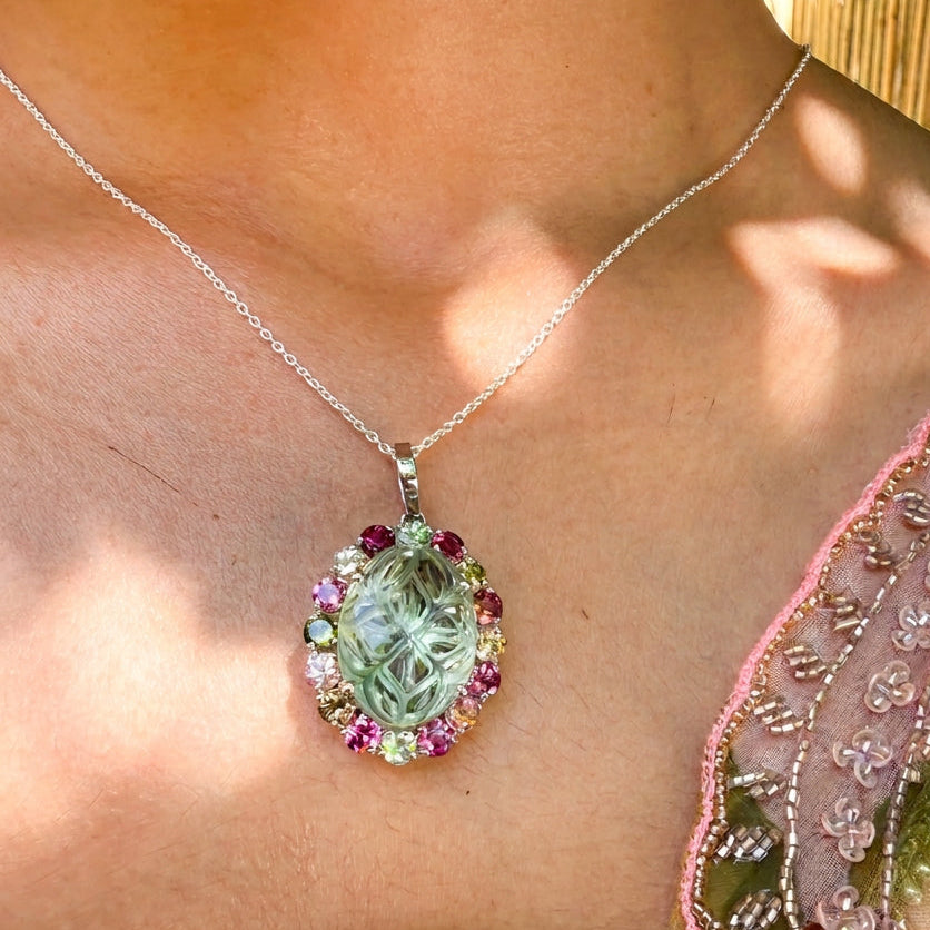 A close-up of a person wearing a silver chain necklace with a teardrop-shaped carved green pendant, accented with pink and yellow gemstones, against a warm outdoor background.