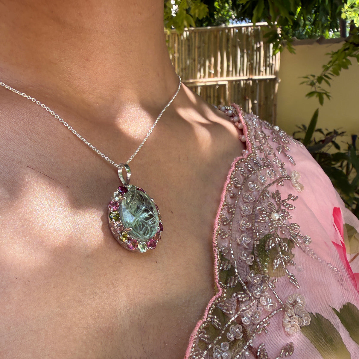 A close-up lifestyle shot of a person wearing a silver chain necklace with a teardrop-shaped pendant. The pendant features a light green central stone surrounded by a halo of pink gemstones and a silver border, set against a background of a person's skin and a pink embroidered saree.