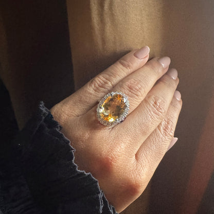 Hand wearing a ring with a large orange gemstone against a neutral background