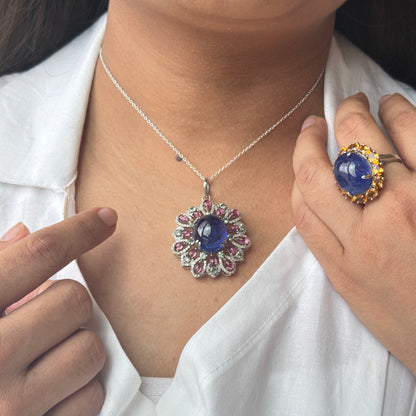 A close-up of a person wearing a silver pendant necklace with a blue cabochon and pink gemstone accents, along with a matching blue ring on a gold band. The person is wearing a white collared shirt.