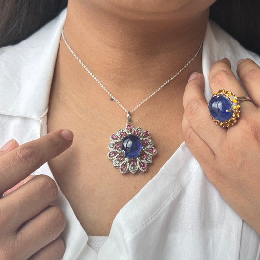 A close-up of a person wearing a silver pendant necklace with a blue cabochon and pink gemstone accents, along with a matching blue ring on a gold band. The person is wearing a white collared shirt.