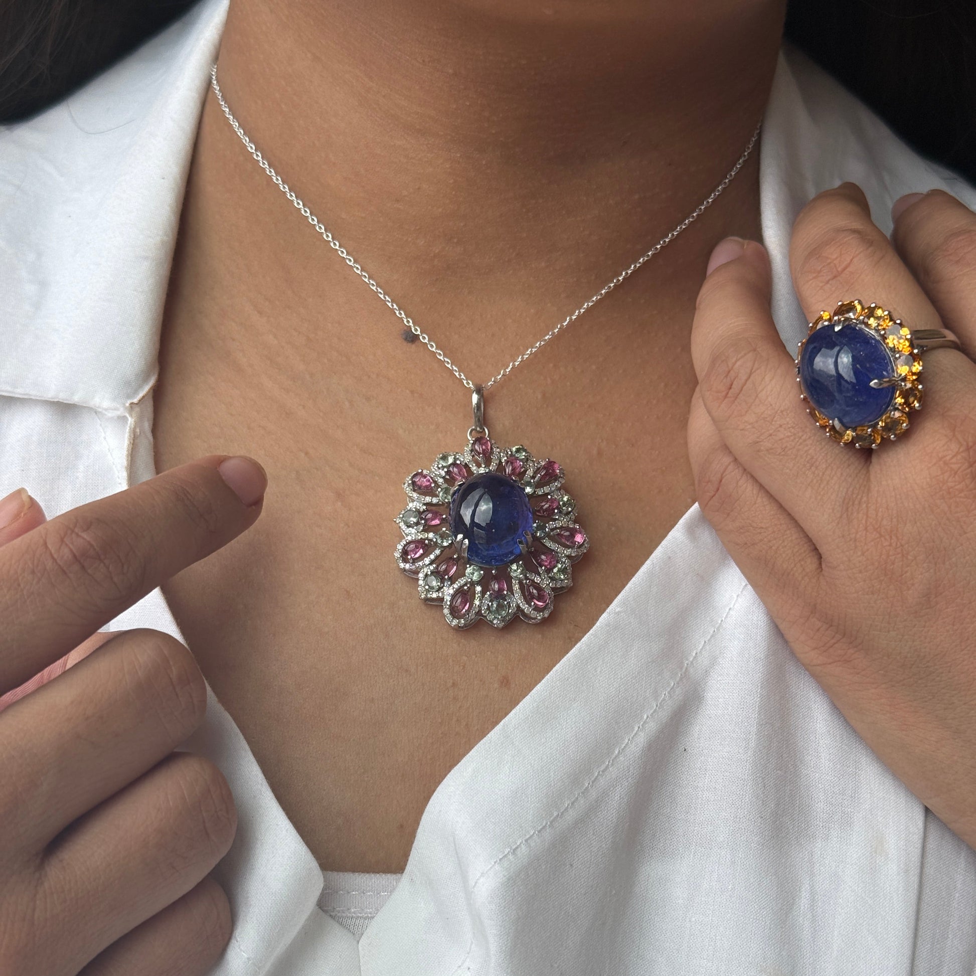 A close-up lifestyle shot of a person wearing a silver necklace with a round blue sapphire pendant, styled with a white shirt. The person is also wearing a matching blue sapphire ring with a gold halo.