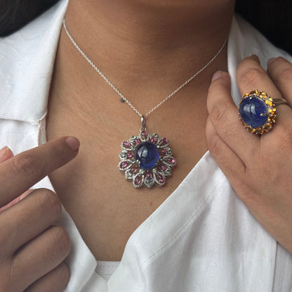 A close-up lifestyle shot of a person wearing a silver necklace with a round blue sapphire pendant, styled with a white shirt. The person is also wearing a matching blue sapphire ring with a gold halo.