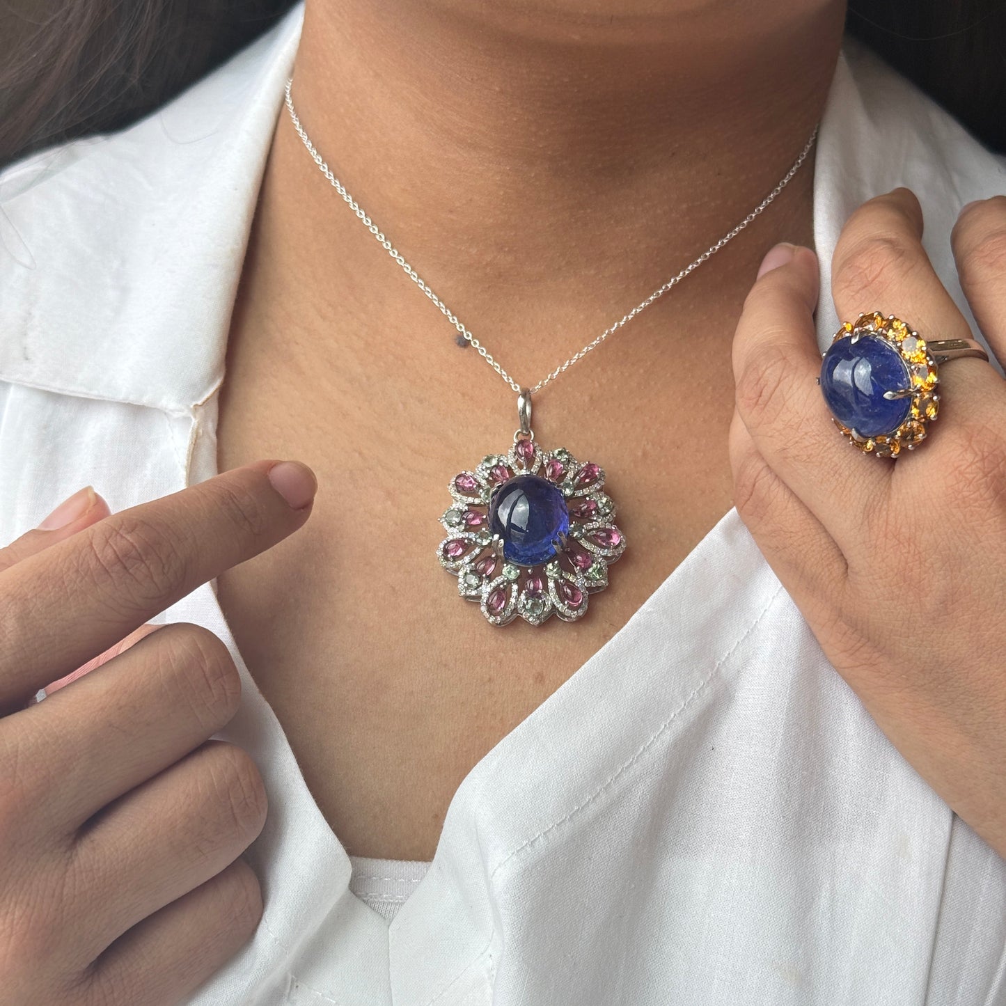 A close-up lifestyle shot of a model wearing a silver pendant necklace with a blue cabochon center and pink gemstone accents, while pointing to the necklace with one hand and wearing a matching ring on the other hand.
