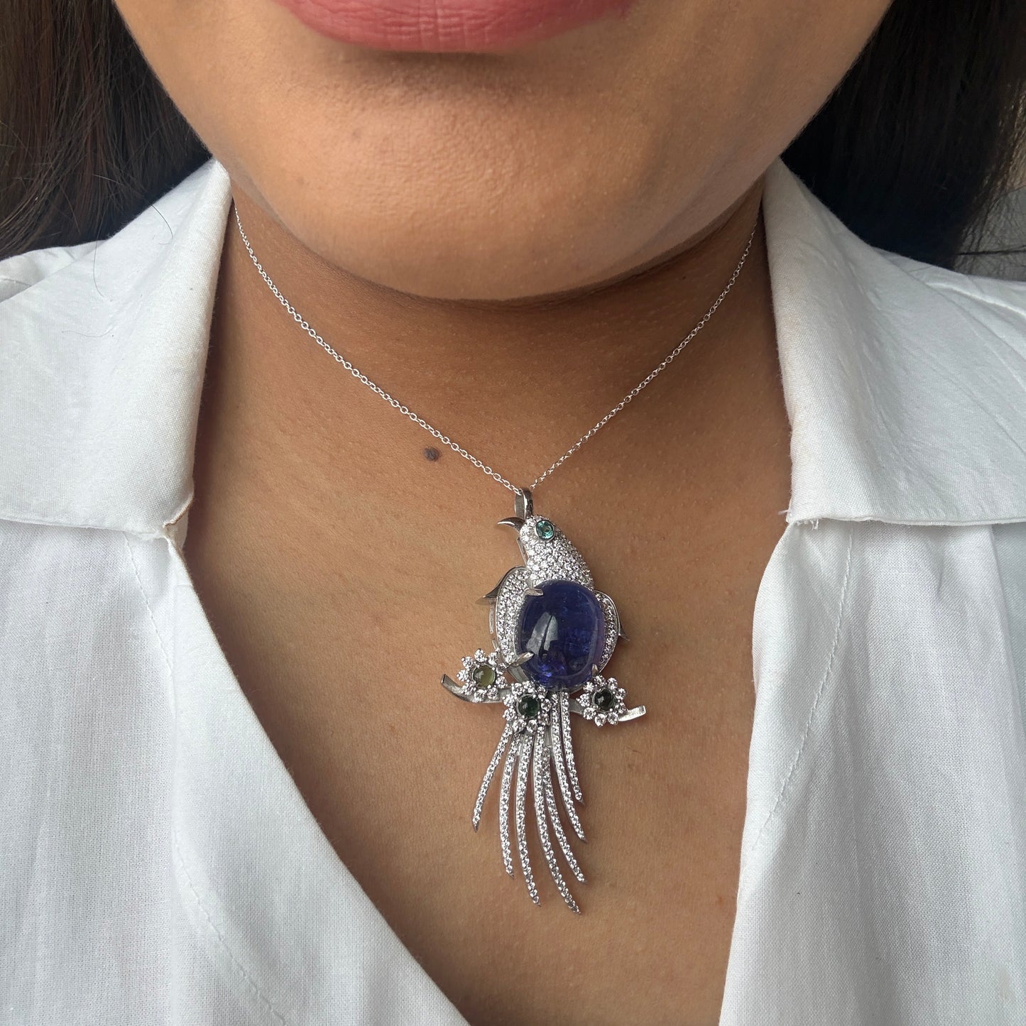 A close-up of a person wearing a silver bird pendant necklace with a dark blue oval center stone and small clear stones, shown against a white collared shirt.