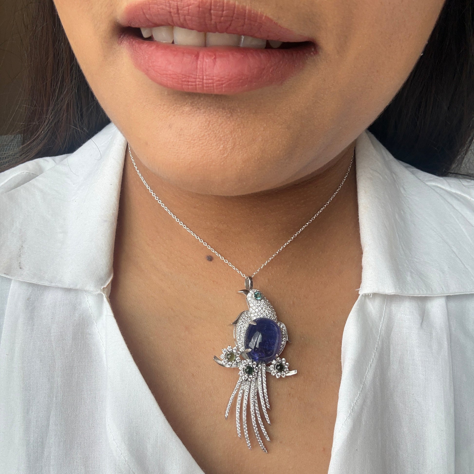 A close-up of a person wearing a silver-toned pendant necklace with a bird design featuring a deep blue oval center stone and small clear stones, styled with a white collared shirt.