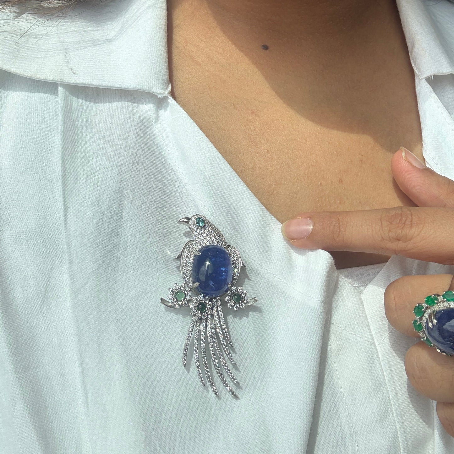 A close-up of a person wearing a silver bird brooch with a blue oval center stone and small green and clear accent stones, pinned to a white collared shirt. A matching ring is also visible on the person's hand.