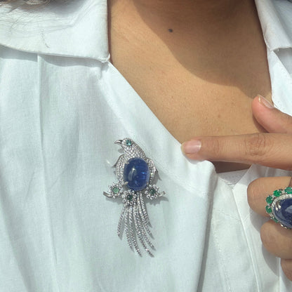 A close-up of a person wearing a silver bird brooch with a blue oval center stone and small green and clear accent stones, pinned to a white collared shirt. A matching ring is also visible on the person's hand.