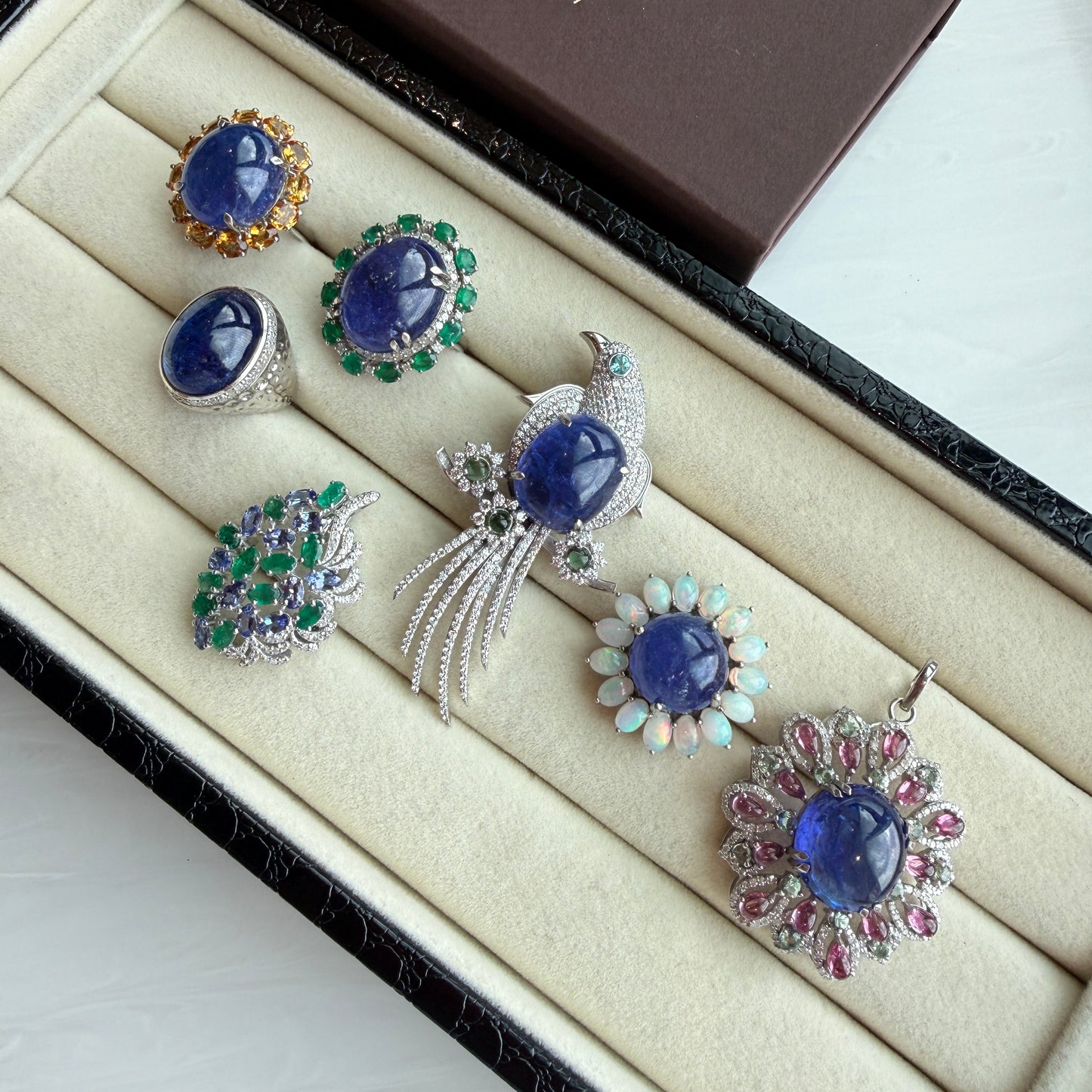 A set of six Indian traditional jewelry pieces, including rings, earrings, and a brooch, displayed in a cream-colored velvet jewelry box. The jewelry features deep blue stones, green emeralds, and silver and gold settings.