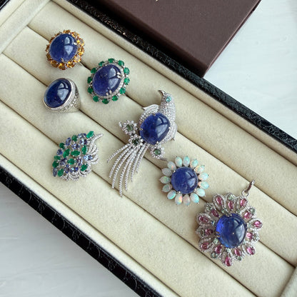 A set of six Indian traditional jewelry pieces, including rings, earrings, and a brooch, displayed in a cream-colored velvet jewelry box. The jewelry features deep blue stones, green emeralds, and silver and gold settings.
