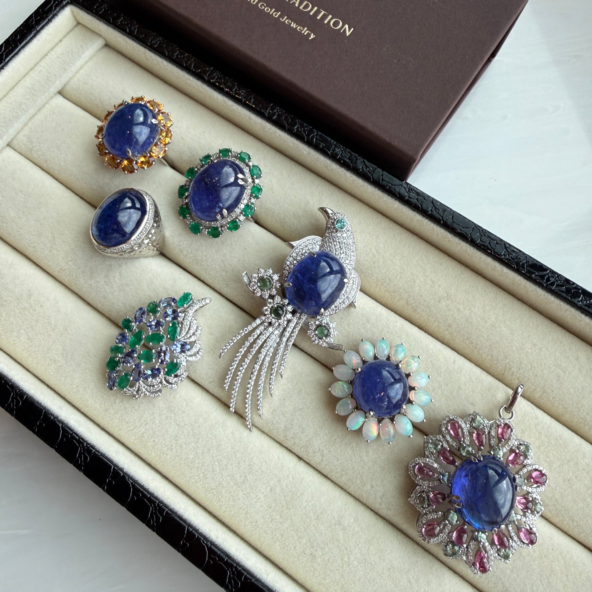 A luxury jewelry set displayed in an open cream-colored velvet box next to its dark brown presentation box. The jewelry collection features multiple pieces with deep blue gemstones, including rings, pendants, and earrings, accented with silver, green, pink, and white stones.