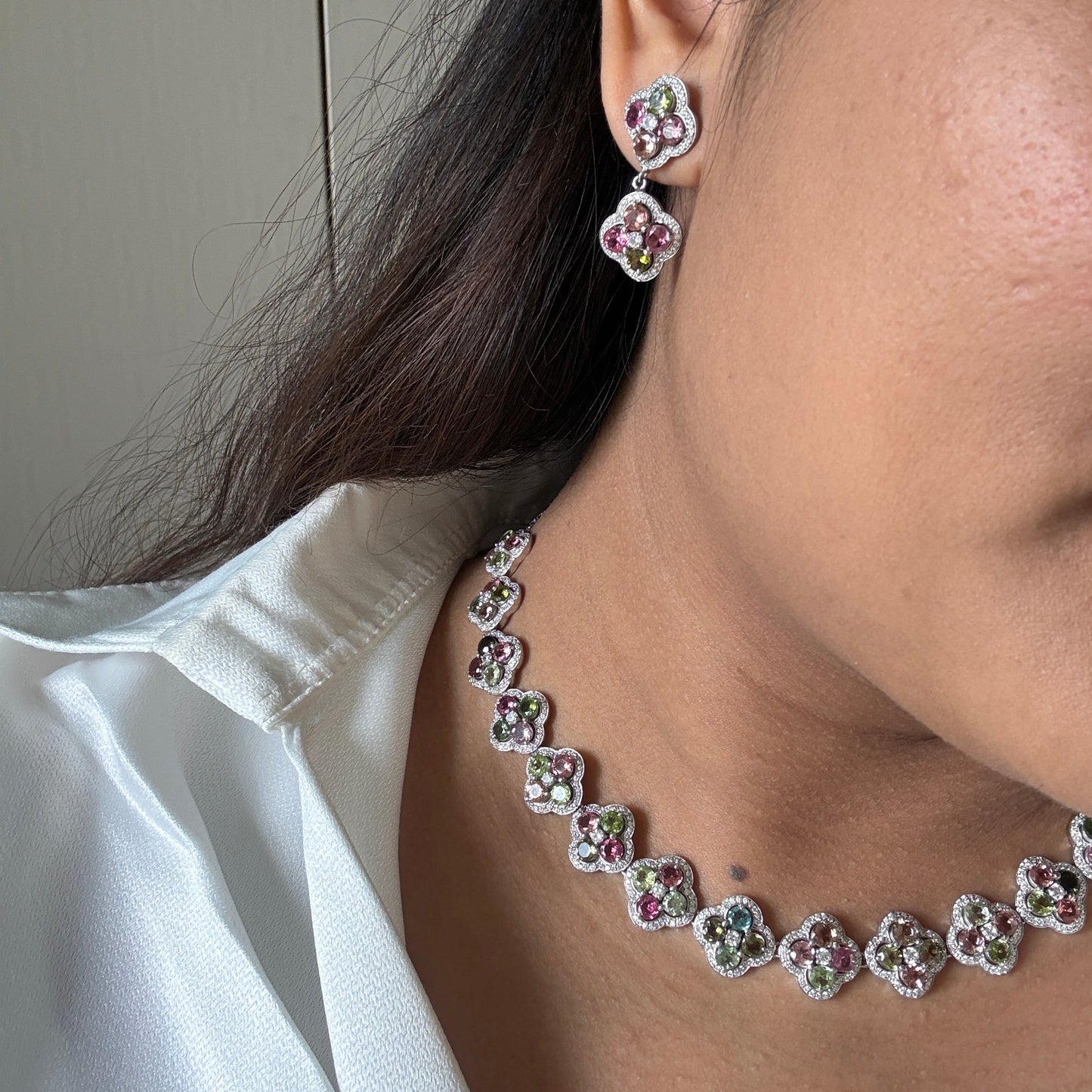 A close-up lifestyle shot of a model wearing a matching silver-toned jewelry set featuring a flower-shaped necklace and matching stud earrings with pink and green gemstone accents.