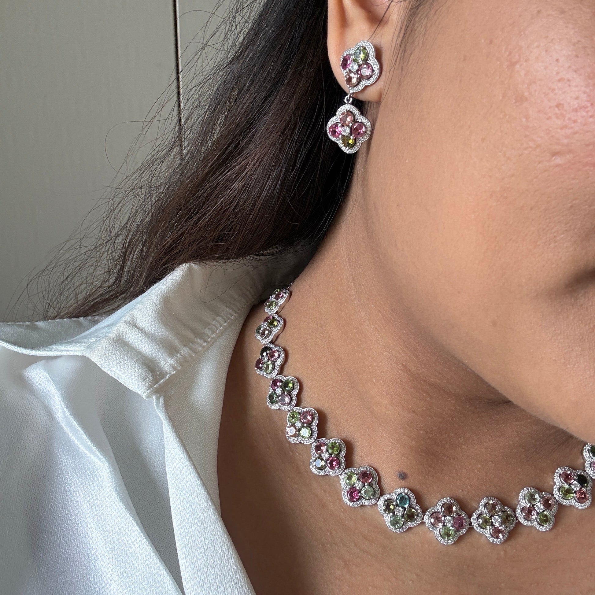 A close-up lifestyle shot of a model wearing a matching silver-toned jewelry set featuring a flower-shaped necklace and matching stud earrings with pink and green gemstone accents.