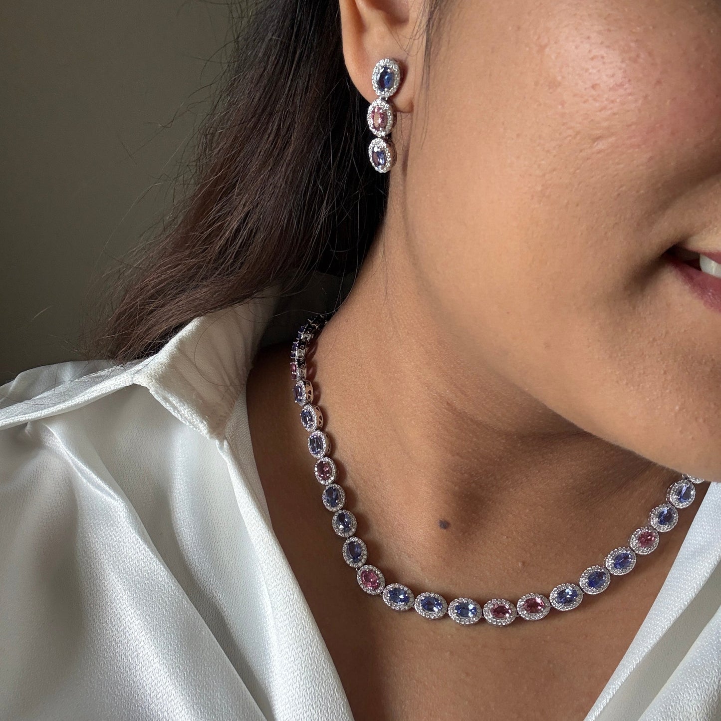A close-up lifestyle shot of a woman wearing a matching jewelry set featuring a silver-toned necklace and drop earring with oval-cut blue and pink gemstones.