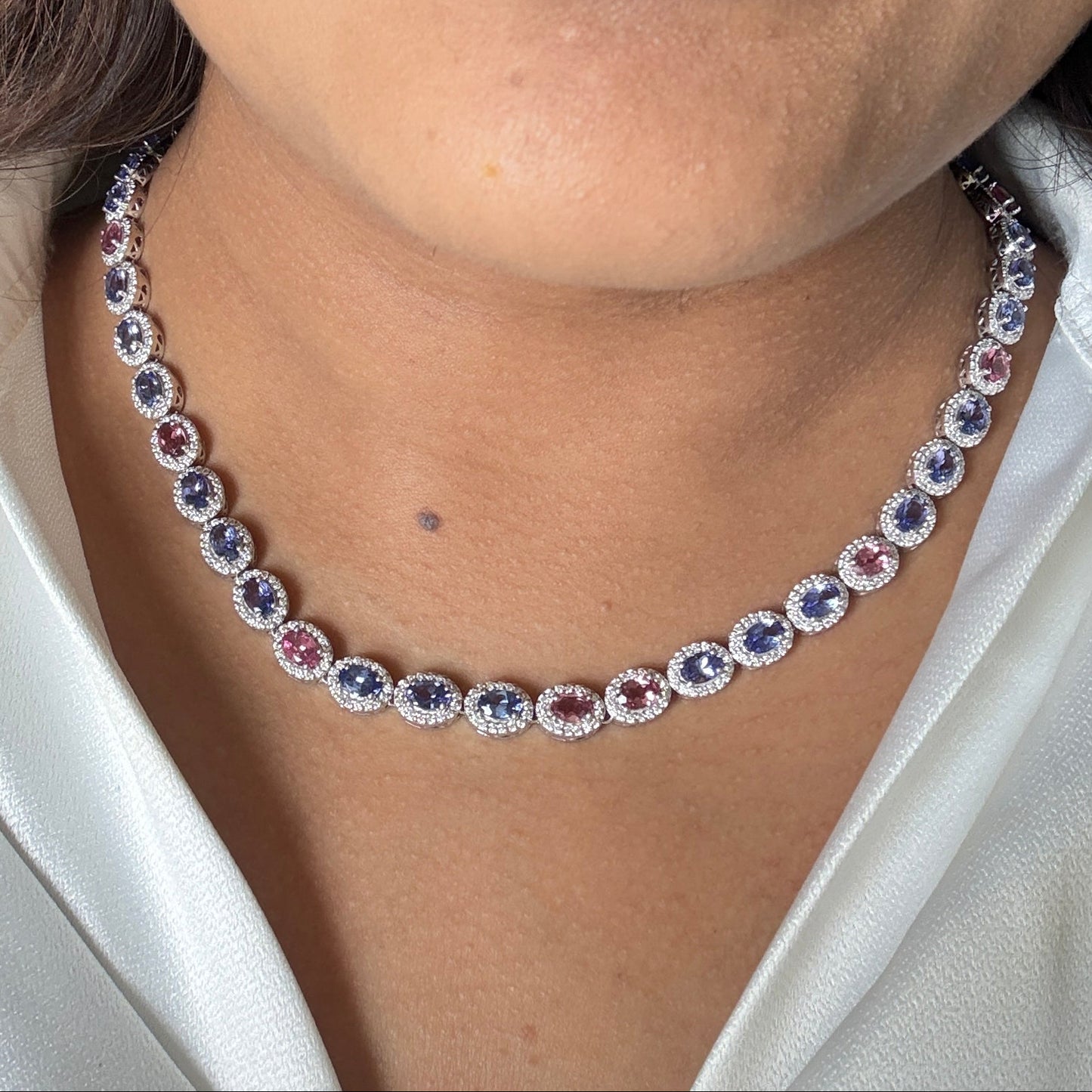 A close-up of a woman wearing a delicate silver-toned necklace with alternating oval-cut blue and clear gemstones, paired with a white satin blouse.