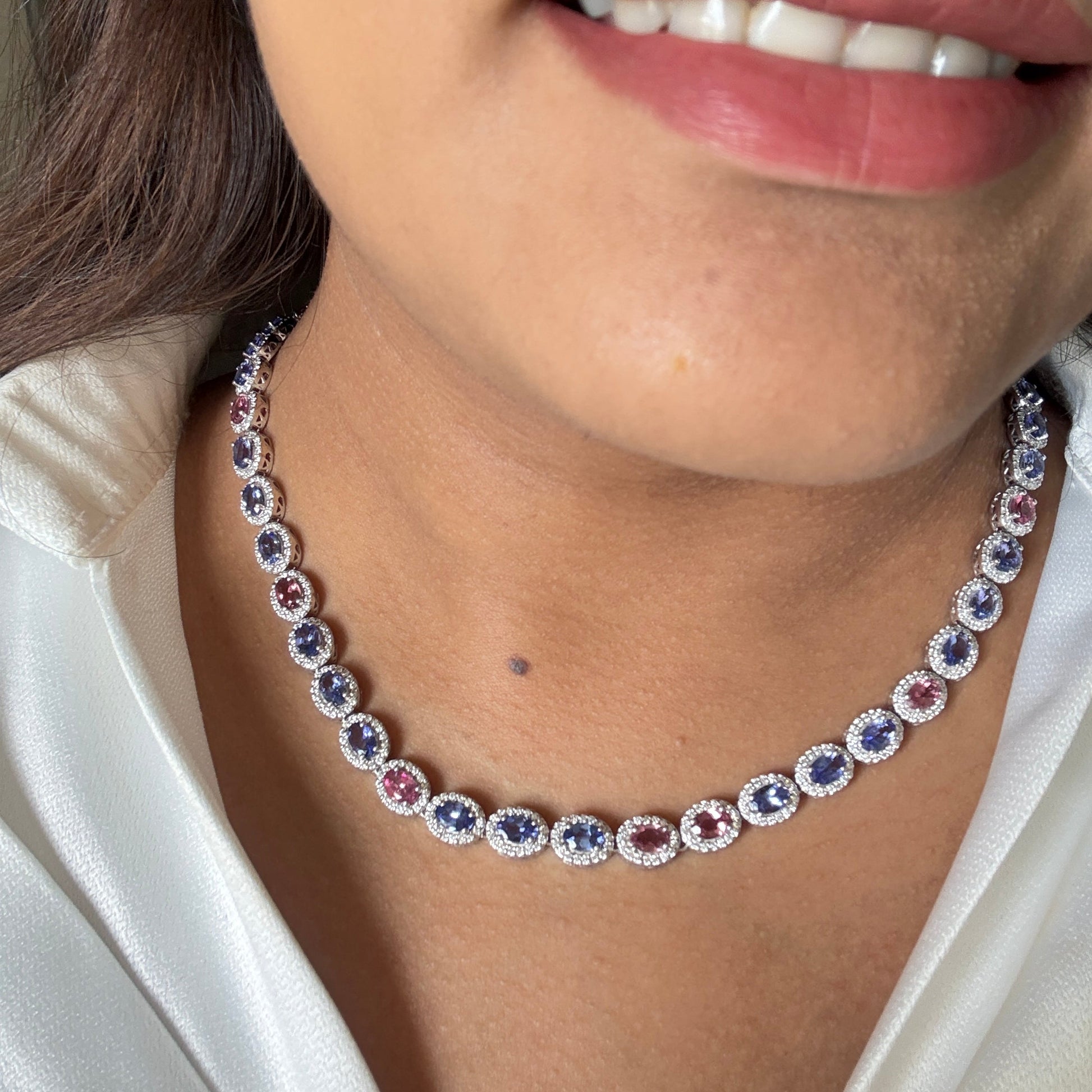 A close-up of a woman wearing a delicate necklace with alternating oval blue and pink gemstones surrounded by small clear stones on a silver-toned chain.