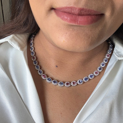 A close-up of a woman wearing a silver-toned tennis necklace with oval blue and pink gemstones, paired with a white satin blouse.