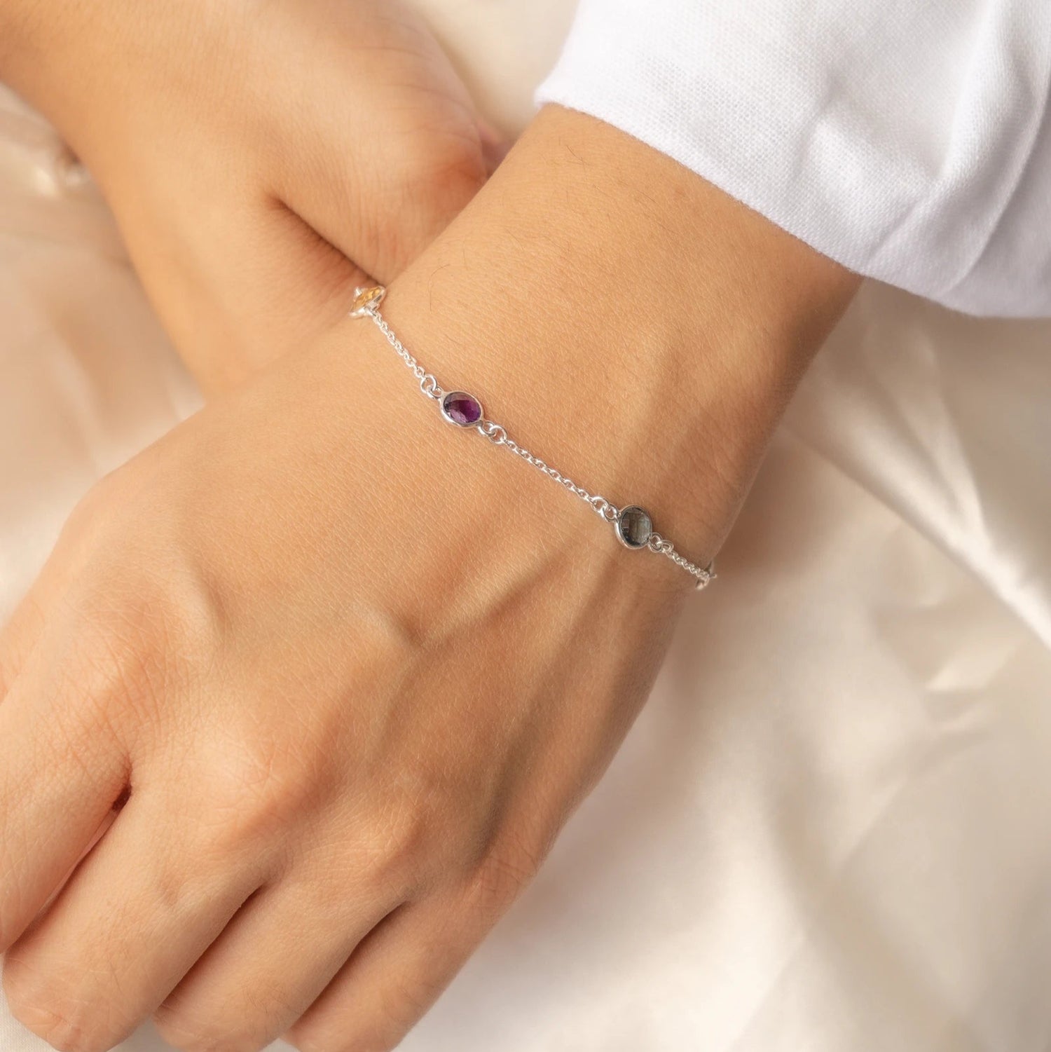 A person's wrist wearing a silver bracelet with round-cut multi-gemstones in purple and green accents.