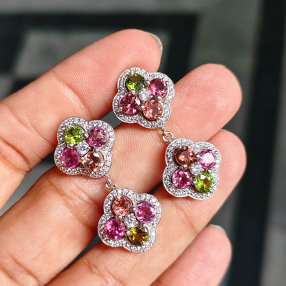 A close-up of a hand holding three matching flower-shaped earrings. Each earring has a silver-toned border with small clear stones and a center cluster of pink, green, and peach gemstones.