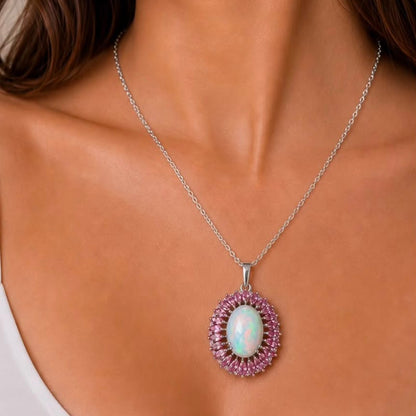 A close-up of a model wearing a silver chain necklace with an oval pendant featuring a white iridescent center stone surrounded by a halo of small pink stones.